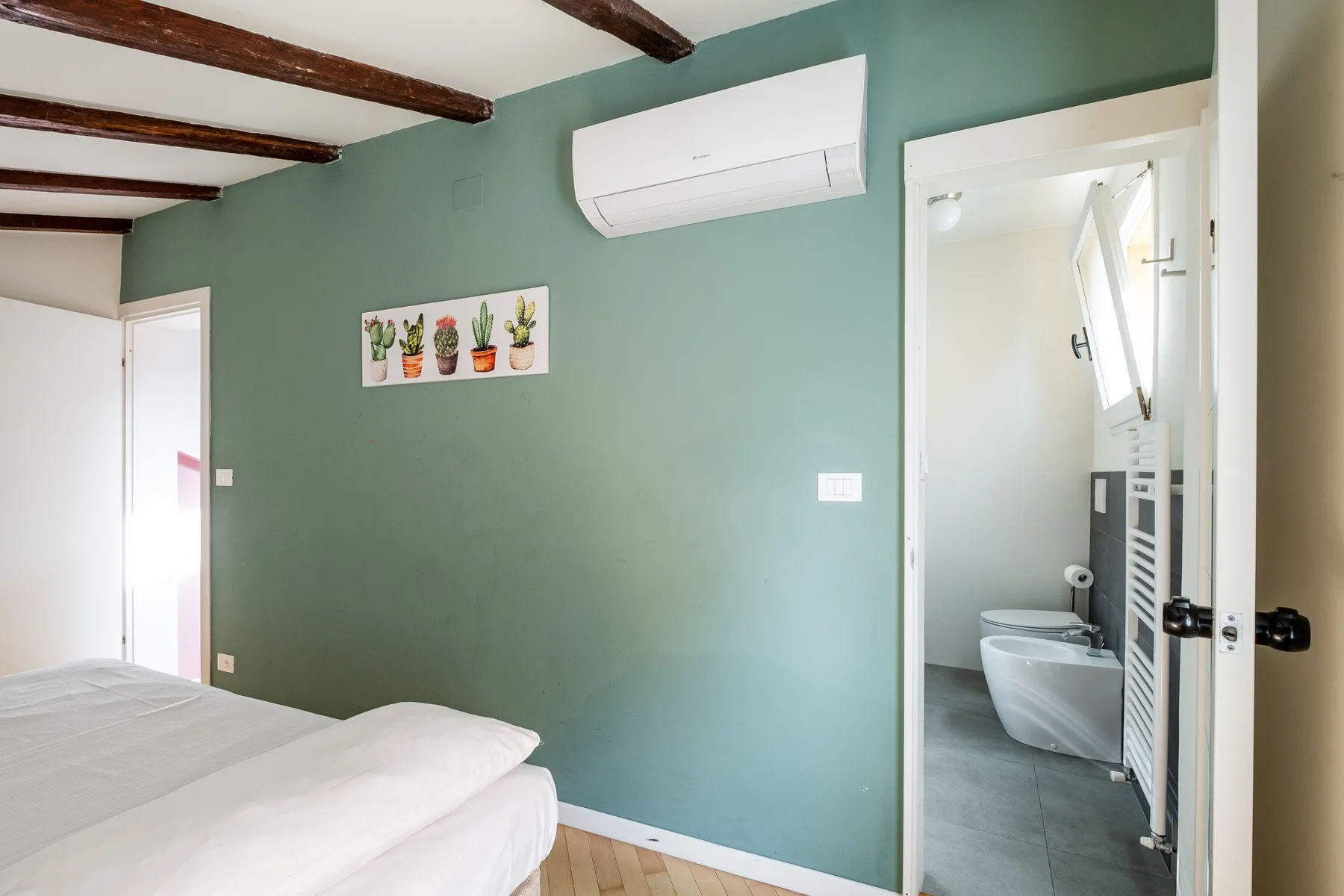 Un dormitorio con ropa de cama blanca y un aire acondicionado de pared junto a un baño abierto.
