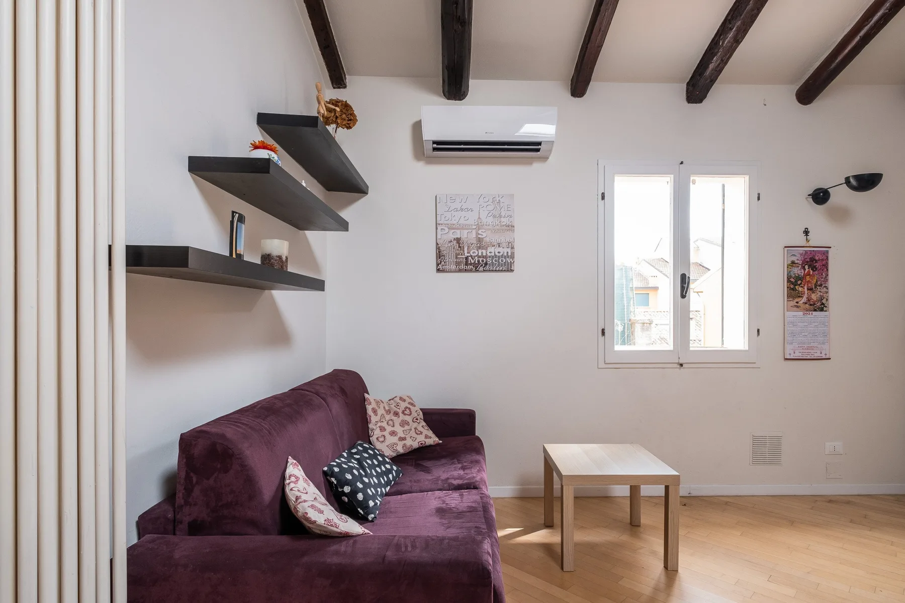 Una sala luminosa con un sofá morado, estantes de pared y una mesa pequeña.