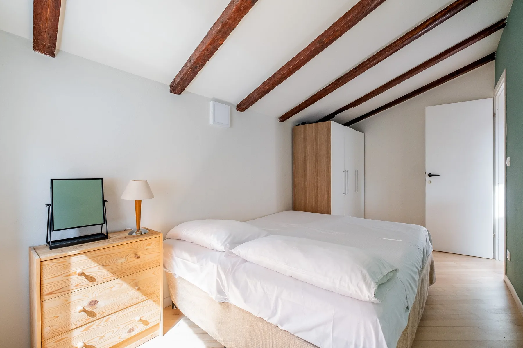 Un dormitorio luminoso con una cama, una cómoda de madera y vigas expuestas en el techo.