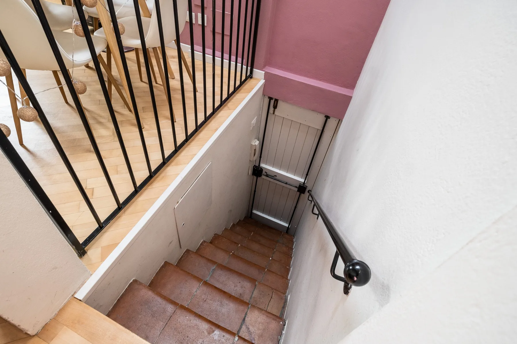 Una escalera de baldosas baja hacia una puerta metálica, con un pasamanos junto a la pared.