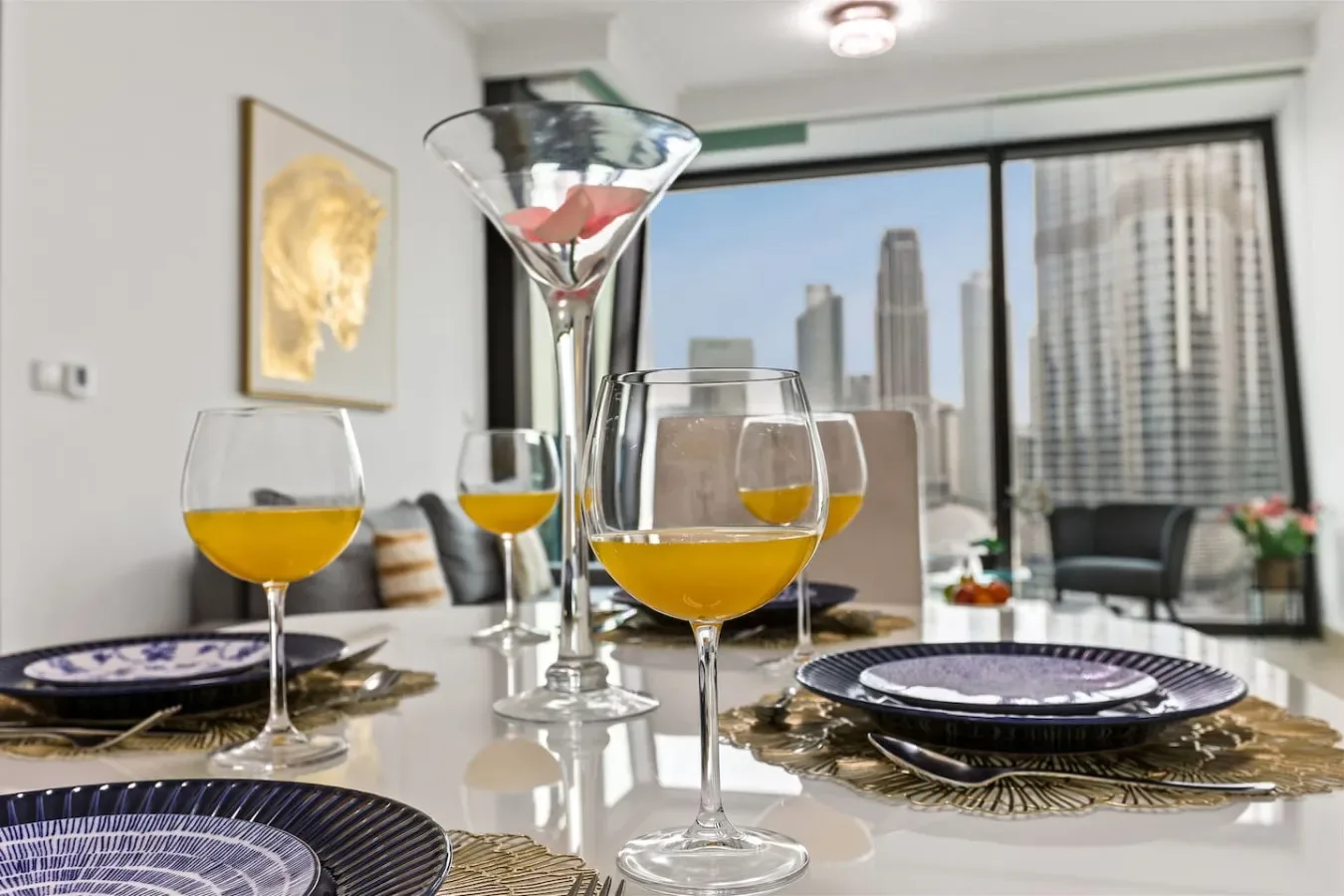 A dining table is set with plates and glasses of orange juice beside a city-view window.