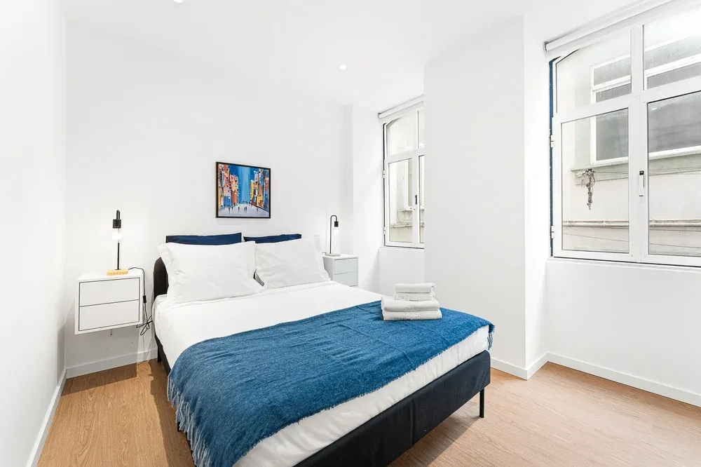 A bright bedroom with a bed covered by a blue blanket and large windows.