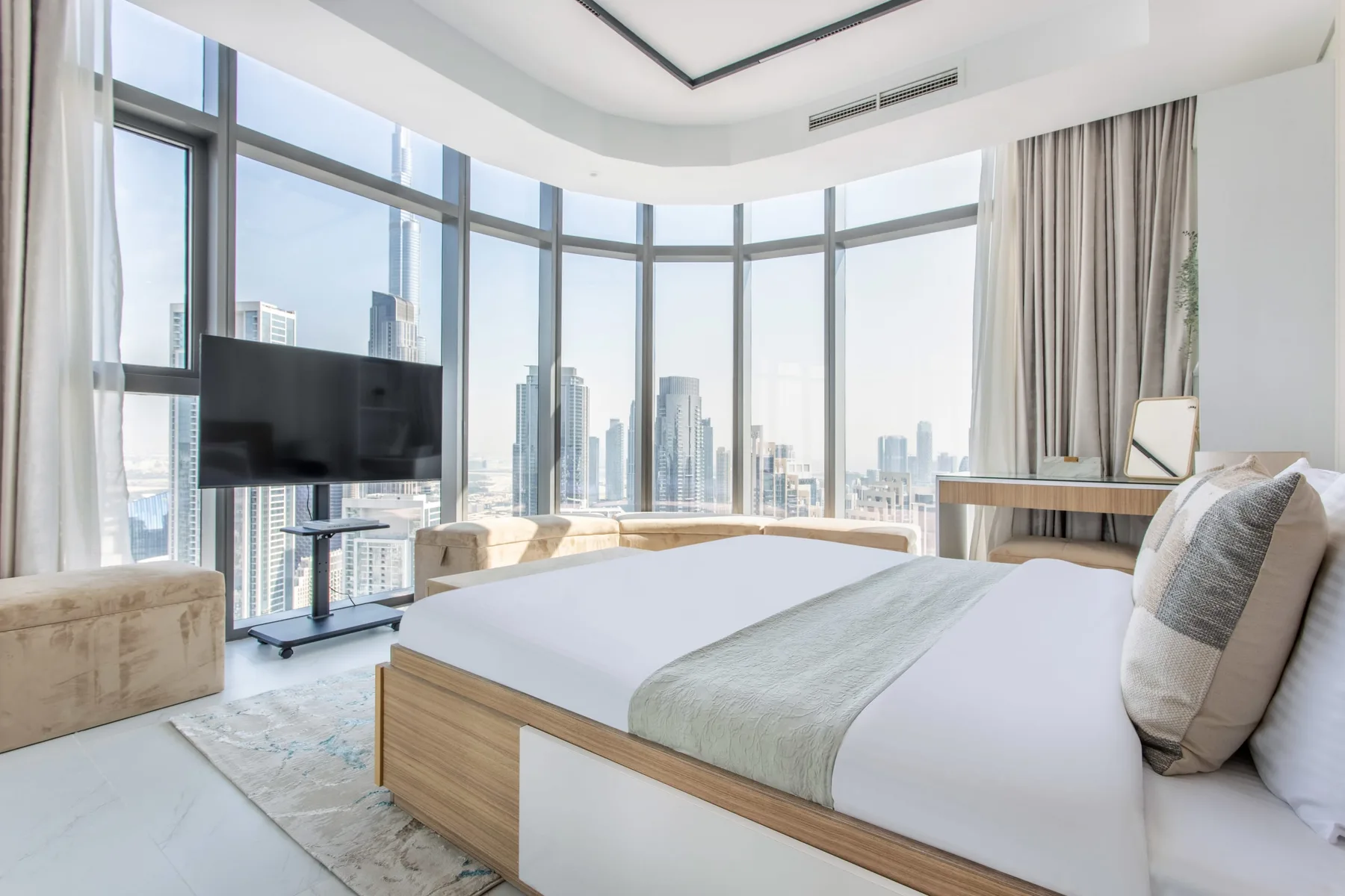 A bright bedroom with a large bed beside floor-to-ceiling windows overlooking city buildings.