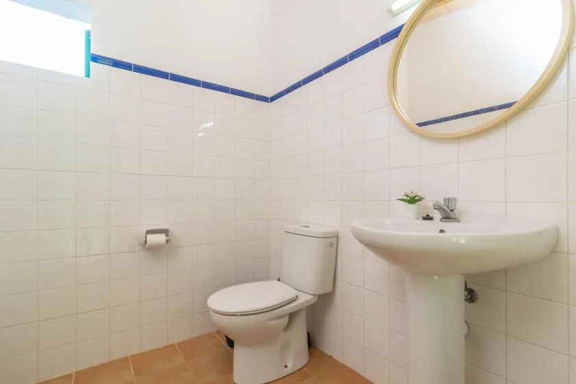 A tiled bathroom with a toilet and pedestal sink beneath a round mirror.