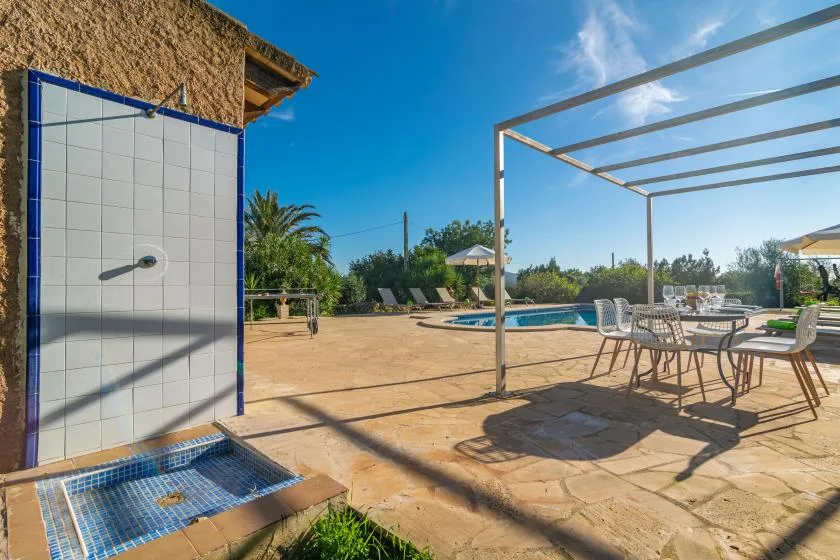 An outdoor pool patio has a tiled shower area and dining seating under a metal frame.