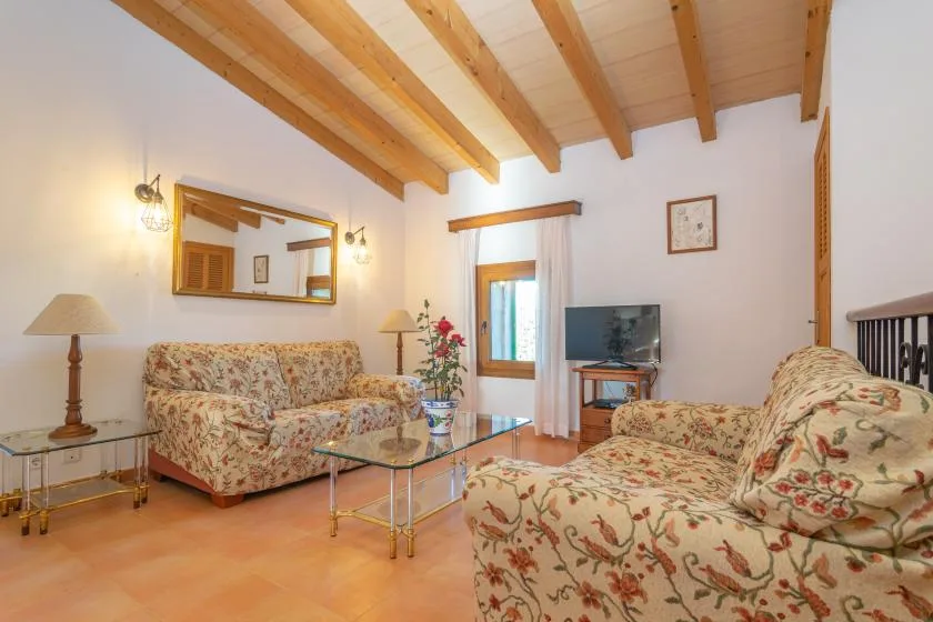A living room with floral seating, glass tables, and exposed wooden ceiling beams.