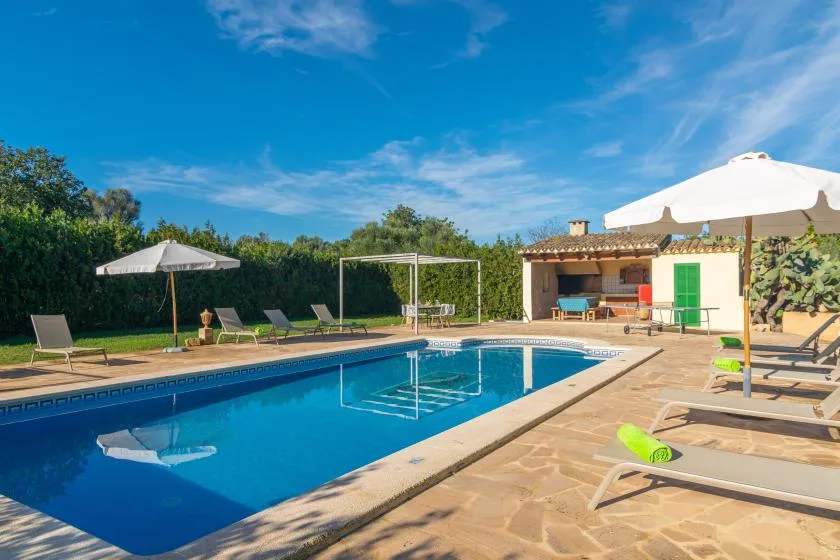 An outdoor swimming pool beside a sunny stone patio with seating and umbrellas.