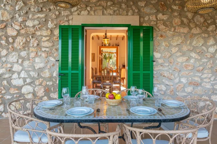 A dining table with place settings sits before open green shutters and a stone wall.