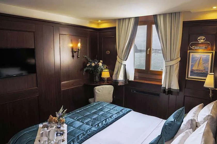 Habitación de hotel revestida de madera con cama, escritorio, ventana y una obra de un velero.