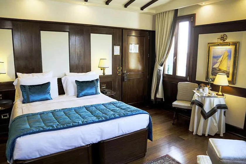 Habitación de hotel con cama, revestimiento de madera, mesa pequeña y una obra de temática náutica.