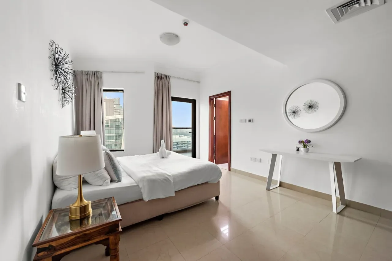 Un dormitorio luminoso con una cama bien tendida, ventanas altas y un escritorio blanco.