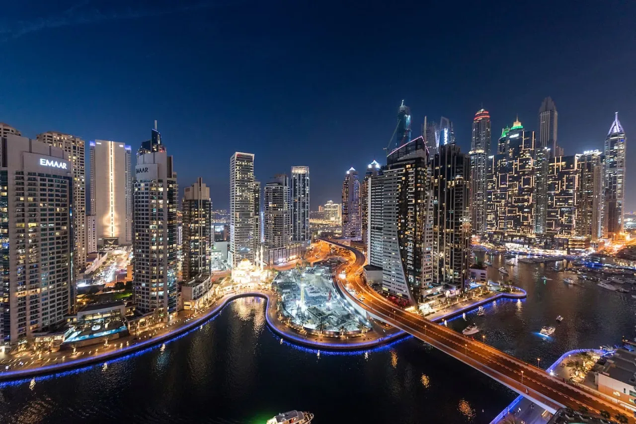 Un horizonte urbano nocturno con edificios altos iluminados y vías junto al agua.