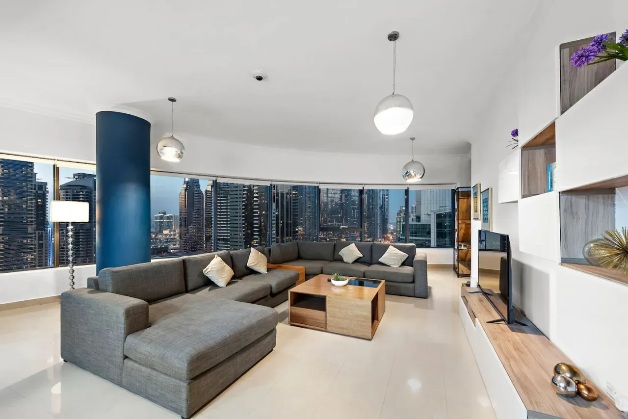 Una amplia sala de estar con asientos grises y grandes ventanales con vistas a edificios de la ciudad.