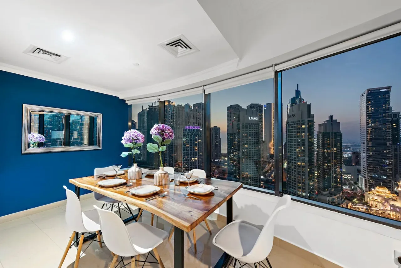 Comedor con una mesa de madera junto a grandes ventanales con vistas a edificios de la ciudad.