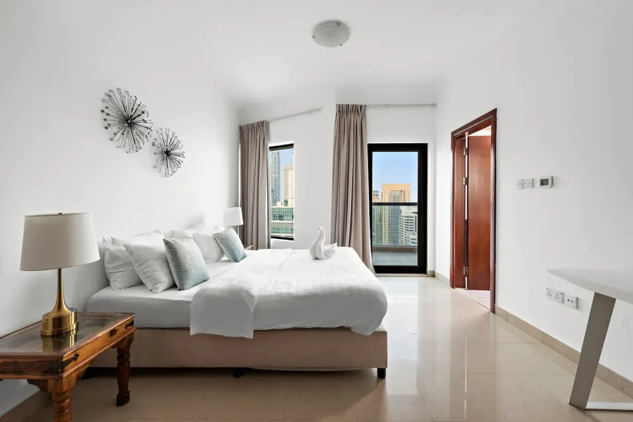 Un dormitorio luminoso con una cama, una lámpara y ventanas altas con vistas a edificios.