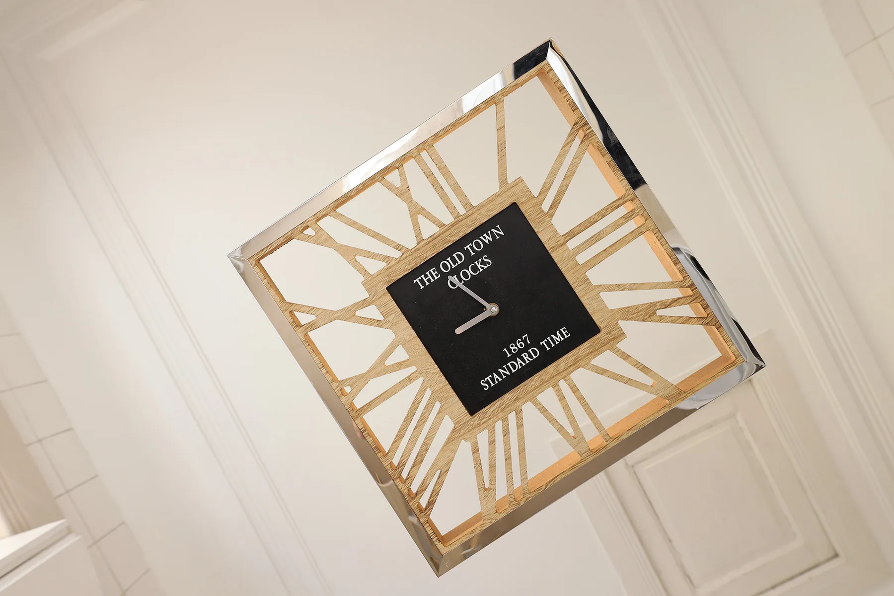 A square wall clock with a gold geometric frame hangs on a white paneled wall.
