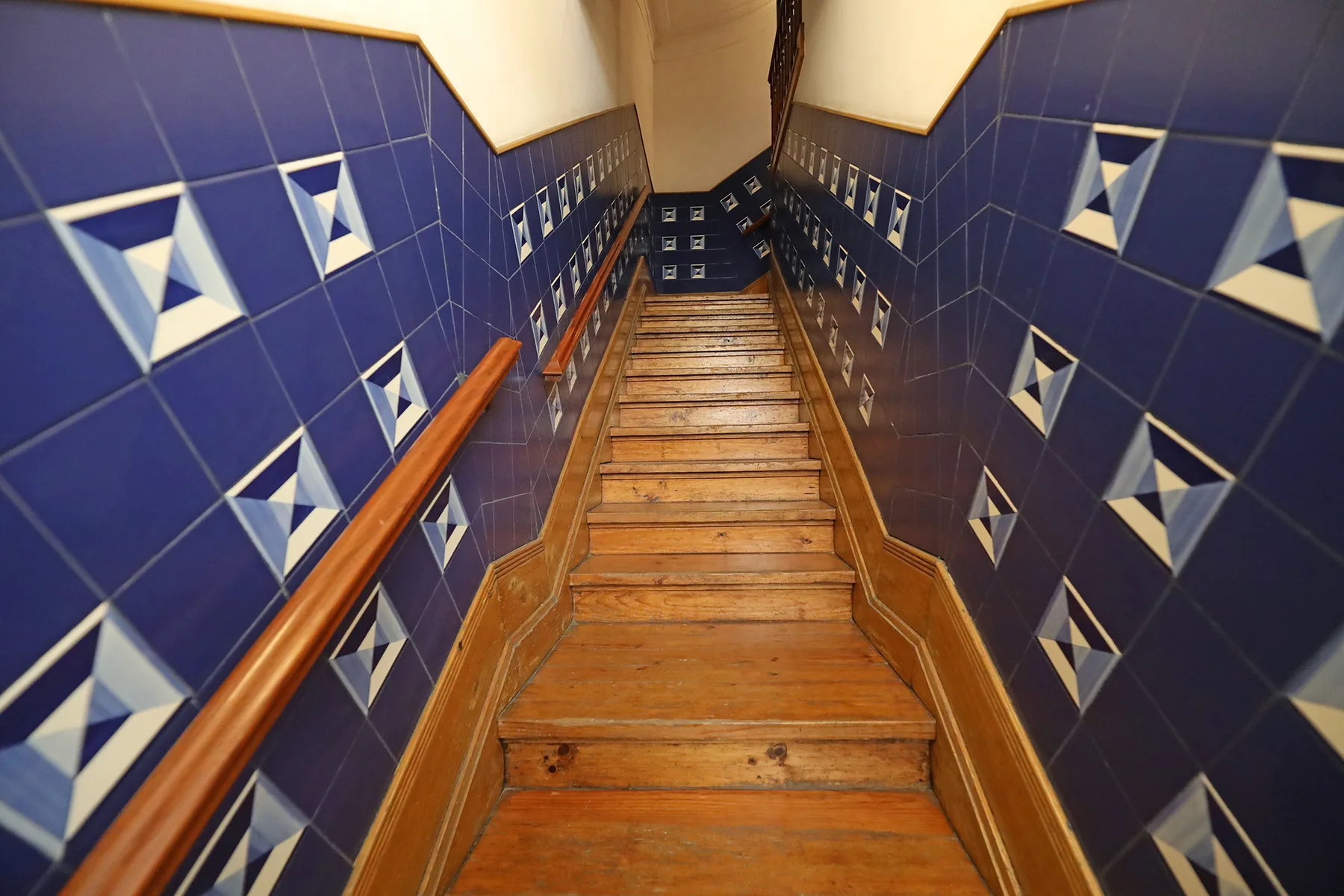 A wooden staircase descends between blue tiled walls with geometric patterns.