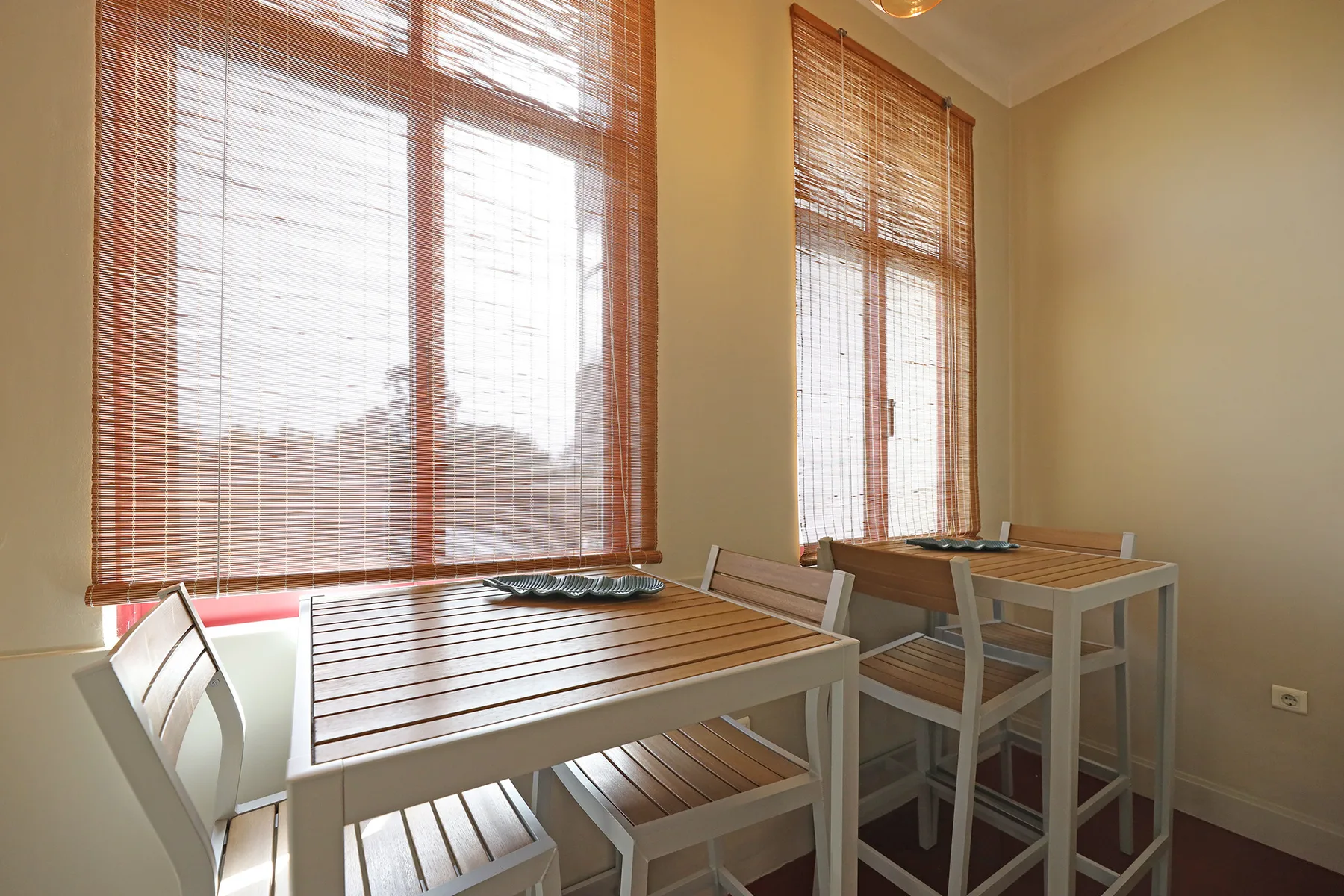 A dining area with tables and chairs beside large windows with woven blinds.