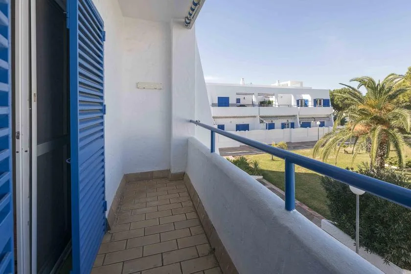 A narrow balcony with blue shutters overlooks neighboring buildings and palm trees.