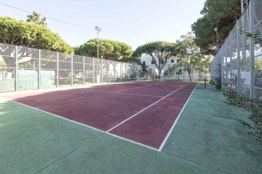 An outdoor tennis court is enclosed by tall fencing and surrounded by trees.