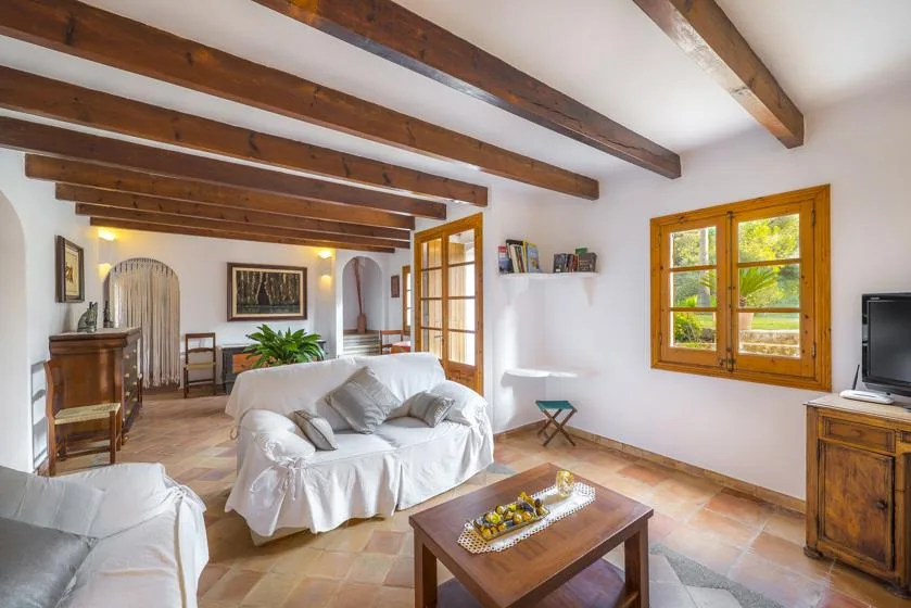 A bright living room with white seating, exposed wooden beams, and a tiled floor.