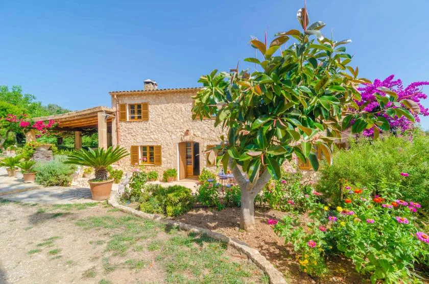 A stone house stands among flowering garden beds and leafy trees.