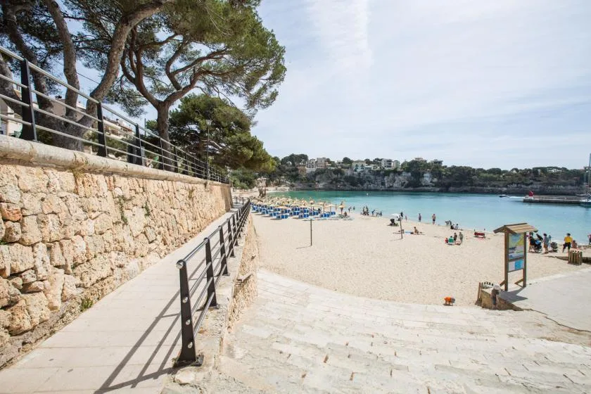 A sandy beach beside a calm bay, with a walkway along a stone wall.