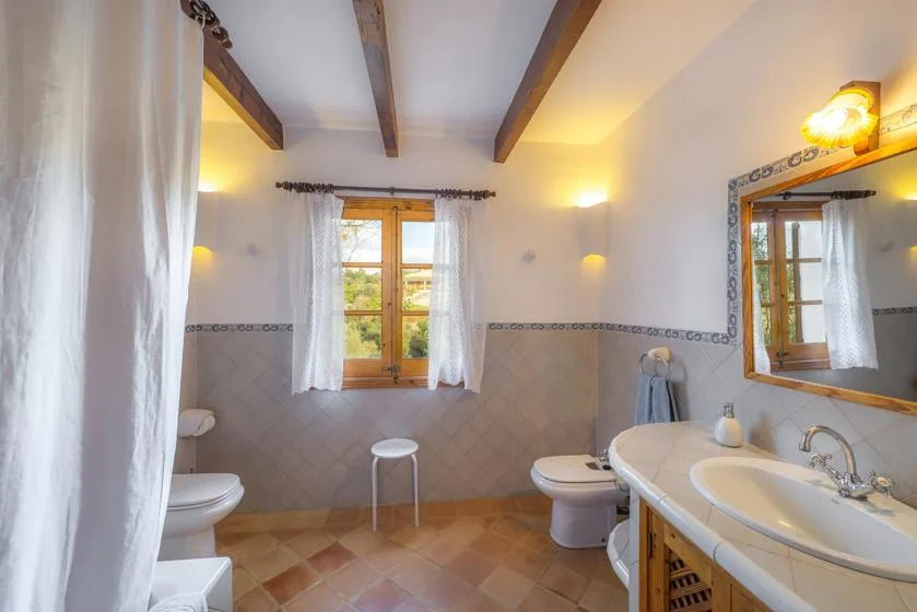 A bright bathroom with a bathtub, toilet, and a window dressed with sheer curtains.