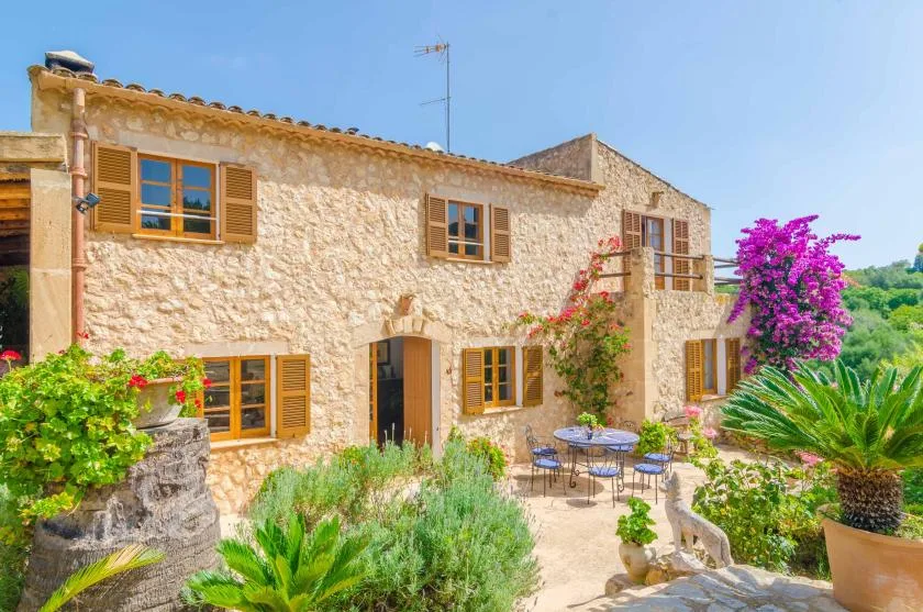 A stone house opens onto a sunny garden patio with flowering plants.