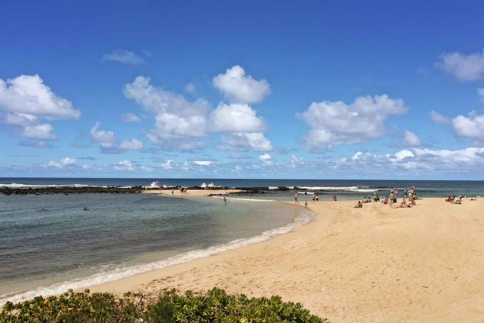 Una playa de arena se curva junto a aguas costeras poco profundas bajo un cielo parcialmente nublado.