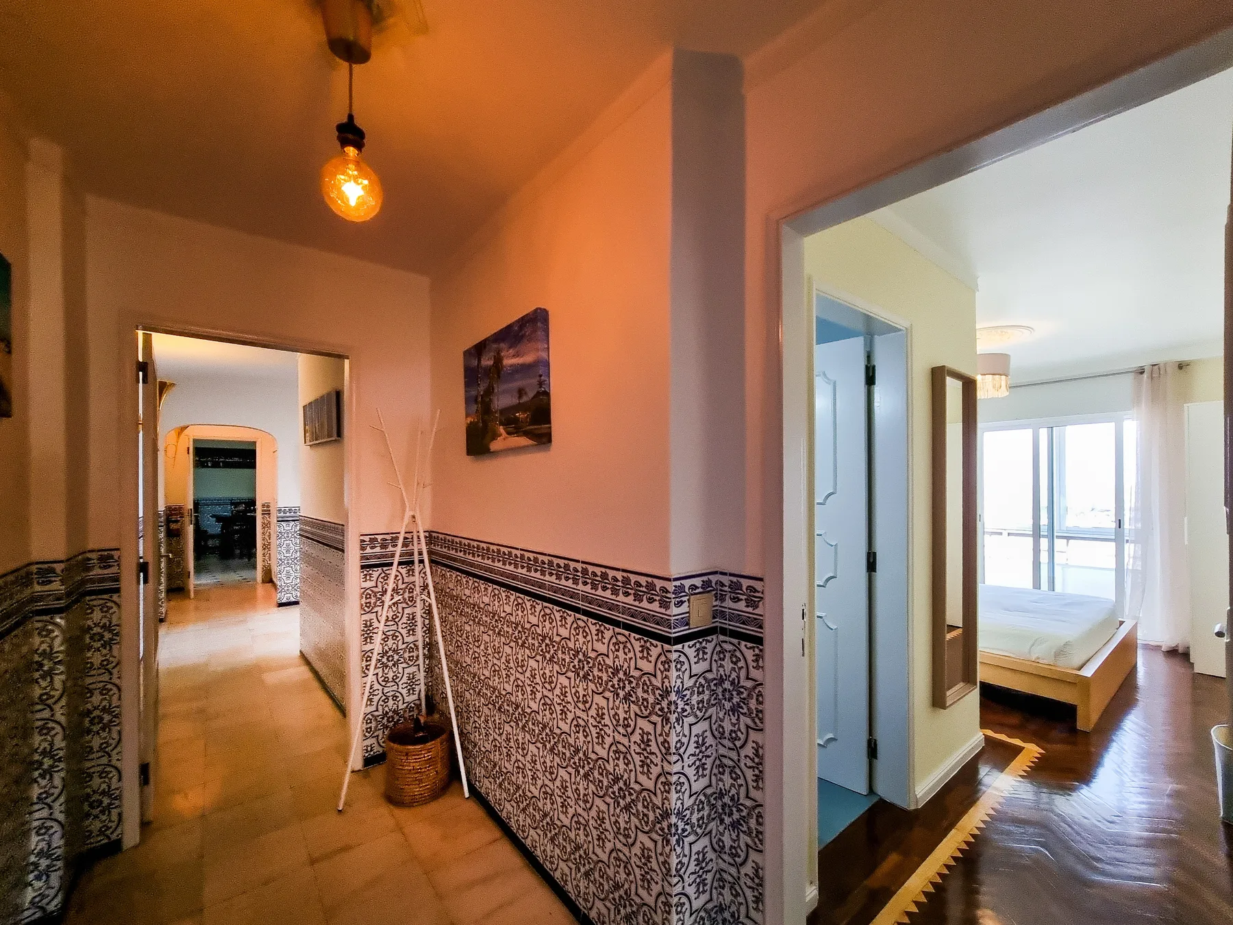 Un pasillo con azulejos da a un dormitorio luminoso con una cama y ventanas grandes.