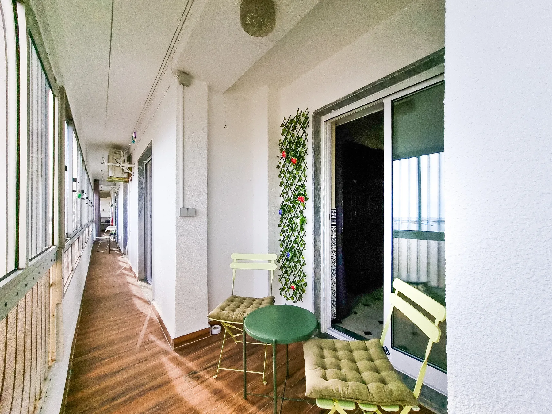 Un largo pasillo de balcón cerrado tiene suelo de madera, ventanas y una pequeña zona de asiento.