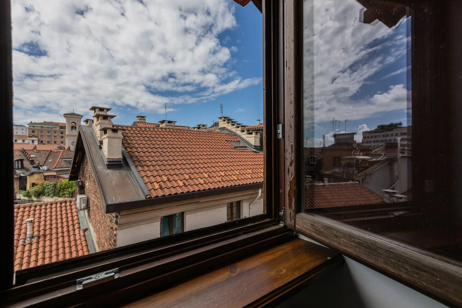 An open window overlooks red-tiled rooftops beneath a cloudy blue sky.