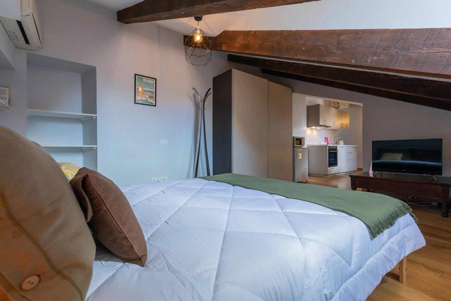 A bedroom with a large bed, exposed ceiling beams, and a compact kitchen area.