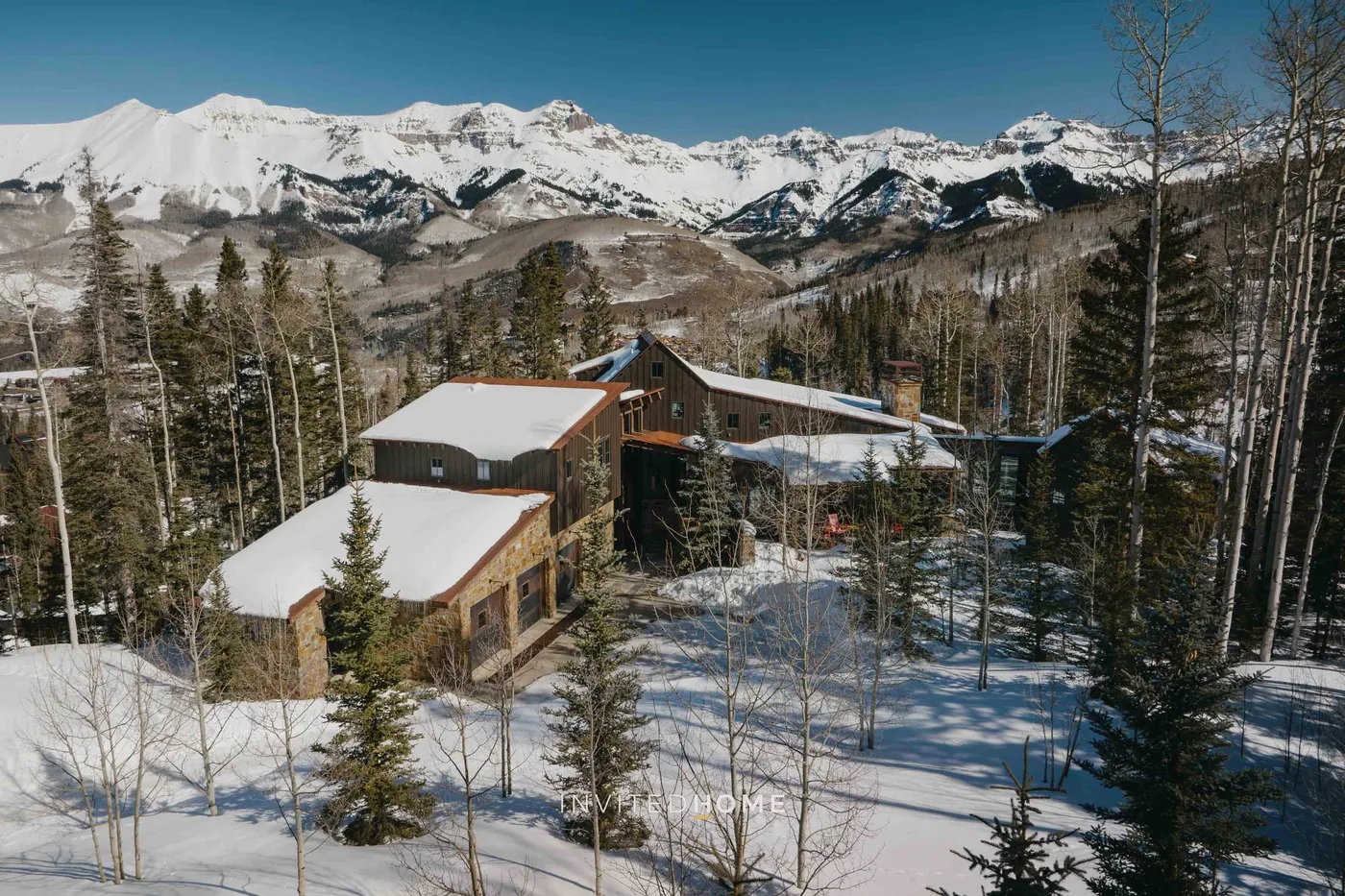 A snow-covered mountain lodge sits among trees with a snowy mountain range in the background.
