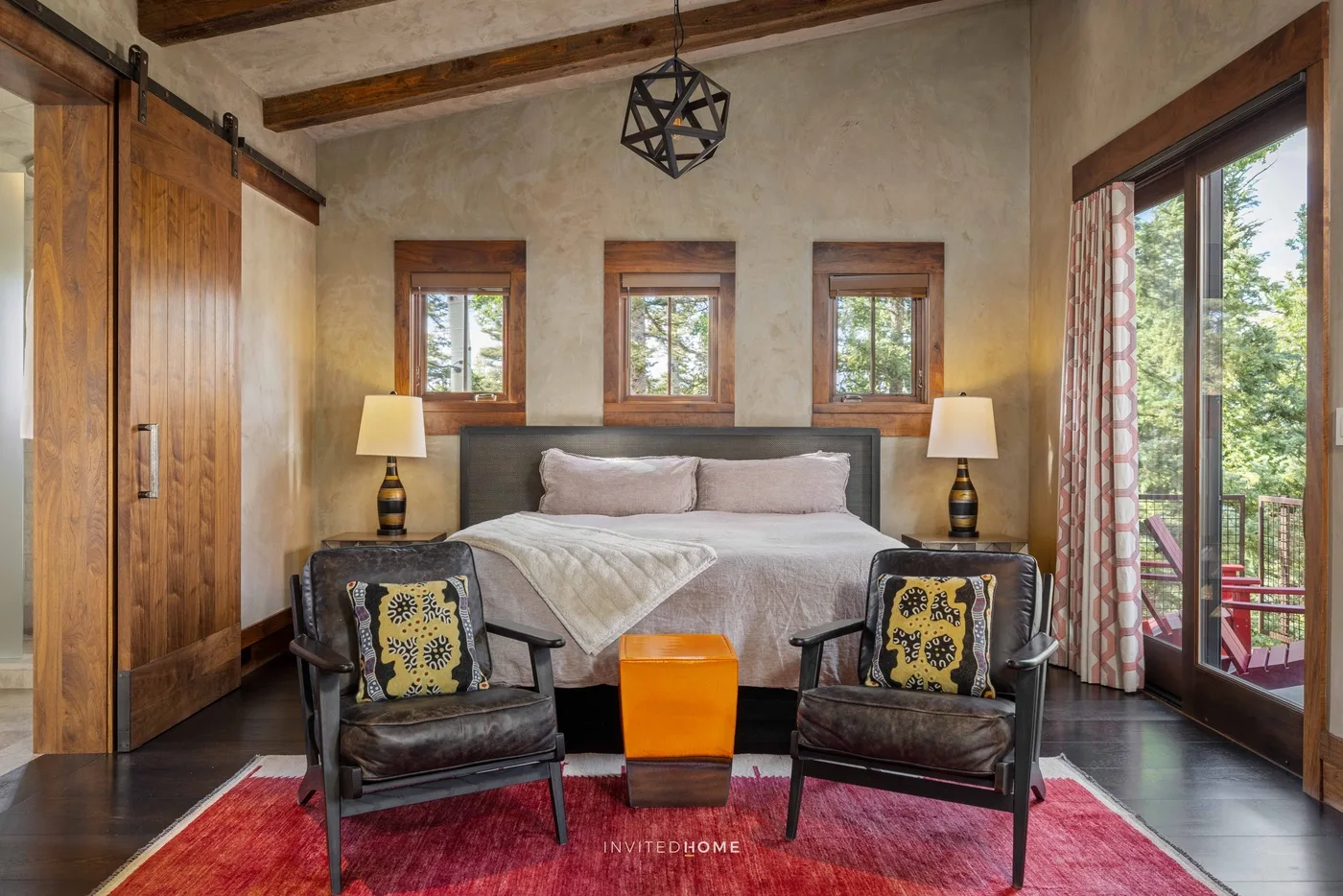 A bedroom with a bed, two armchairs, and windows overlooking greenery.