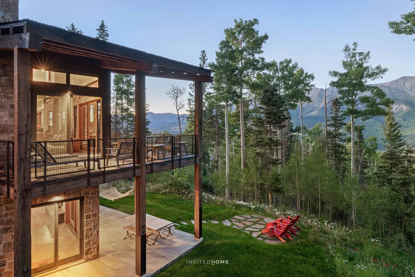 A modern house opens onto a wooded mountain view, with outdoor seating on the deck.