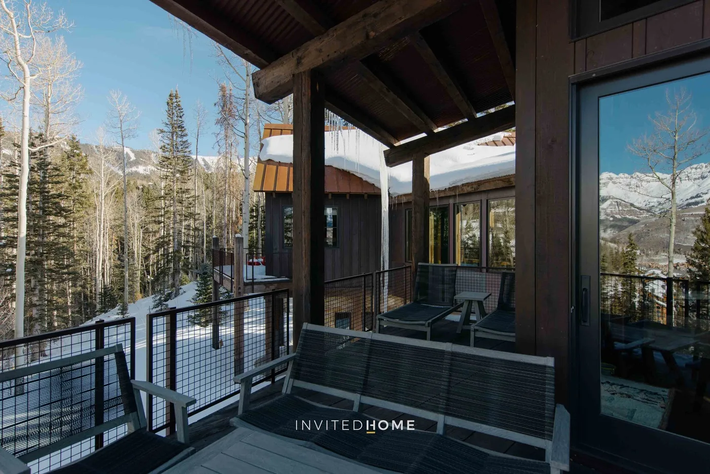 A covered outdoor deck with seating overlooks a snowy, tree-lined mountain landscape.