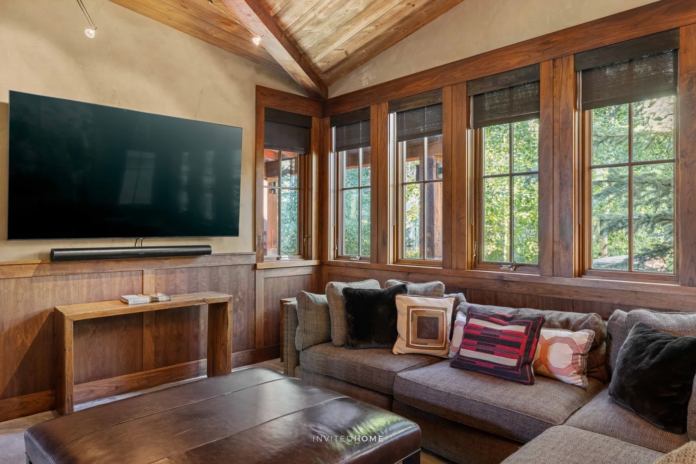 A cozy living room with a television, seating, and large windows overlooking greenery.