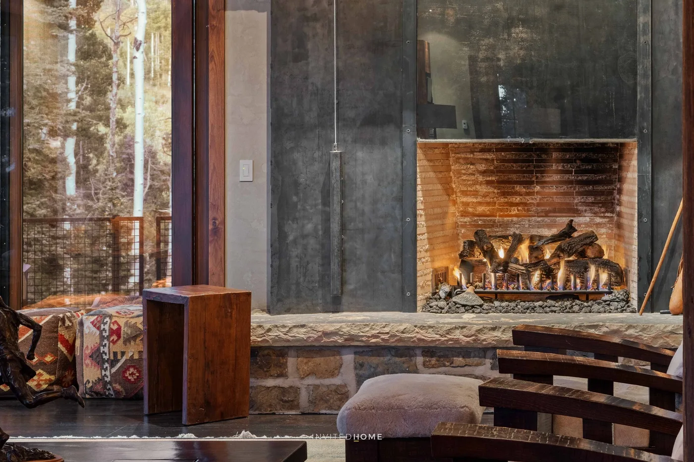 A living room with a large fireplace, low seating, and windows overlooking trees.