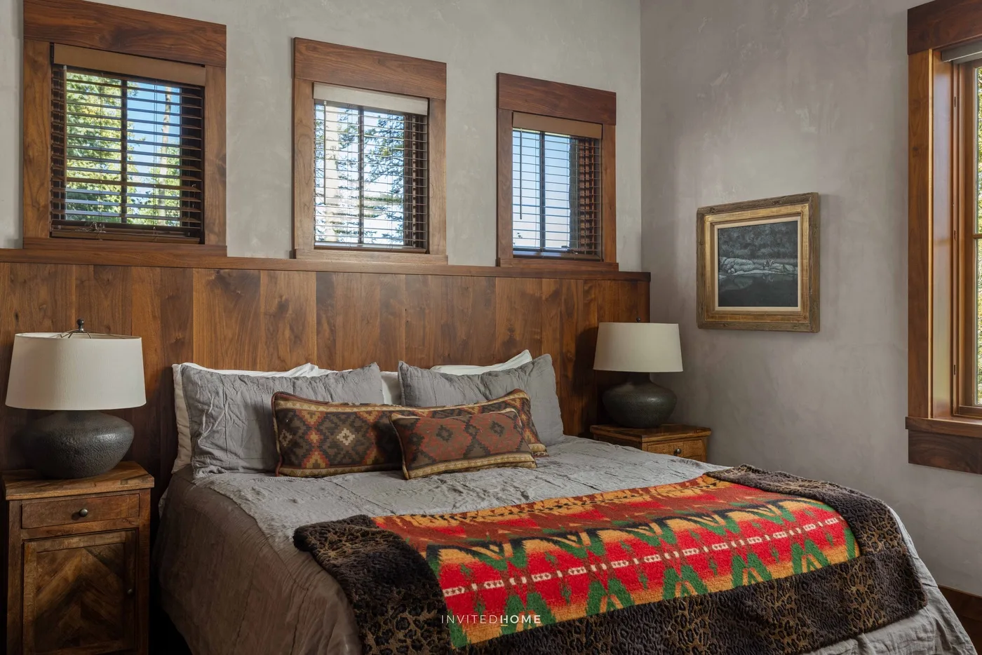 A bedroom with a bed, patterned blanket, and wood-trimmed windows.