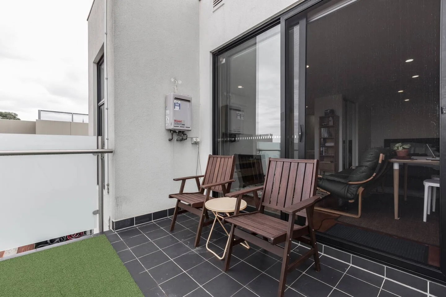 Una zona de estar en el balcón tiene sillas de madera, una mesa pequeña y césped artificial verde.