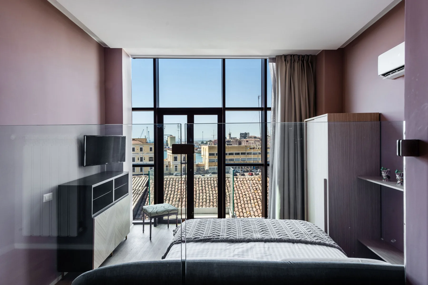 A bedroom with a bed facing a large window overlooking nearby rooftops.