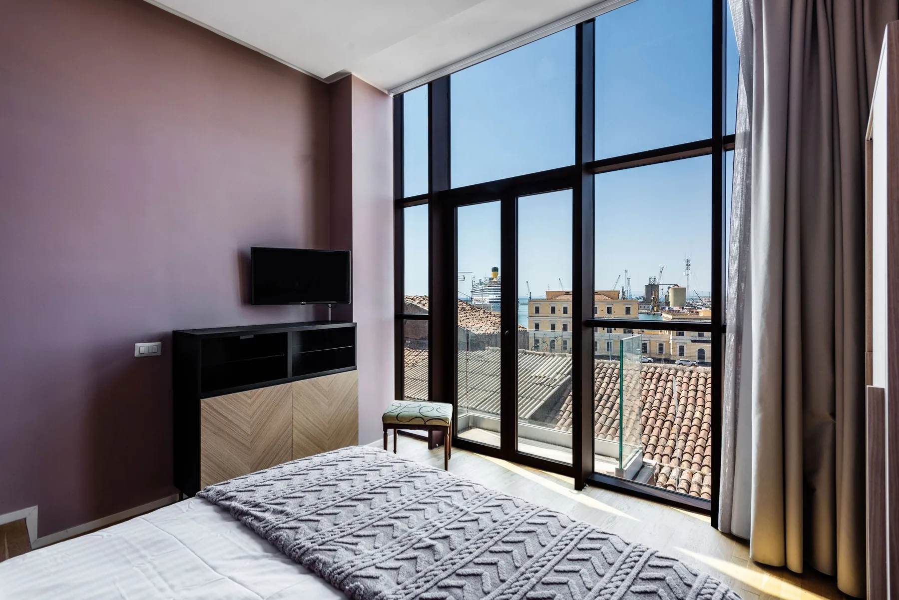 A bedroom with a bed and large windows overlooking nearby rooftops.