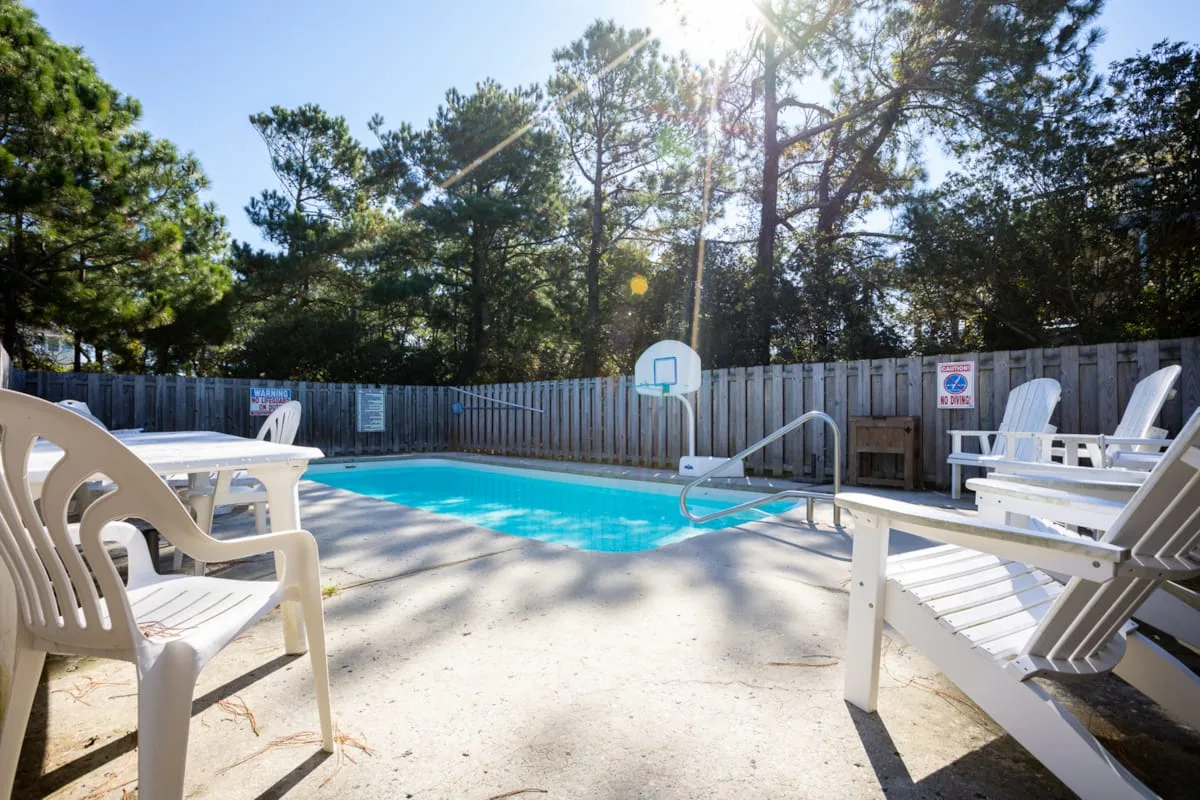 An outdoor swimming pool sits among trees, with lounge chairs and a basketball hoop nearby.