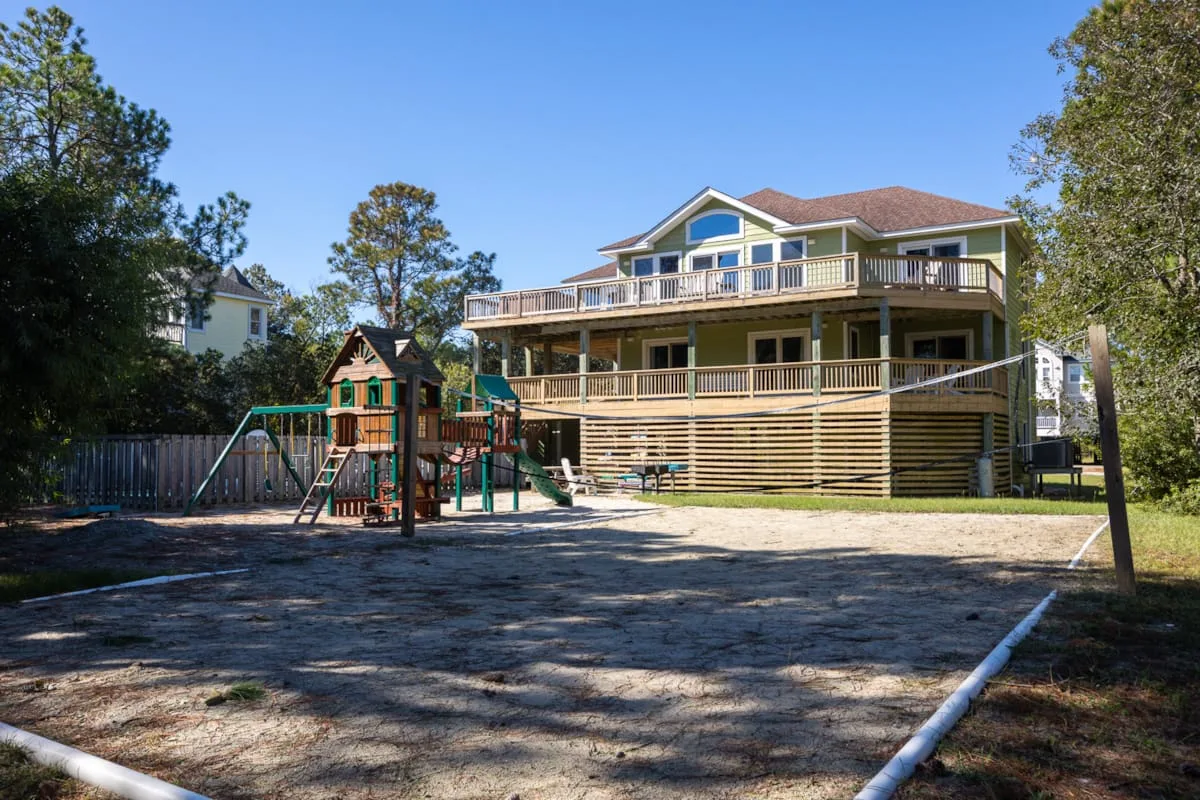 A large house with wraparound decks stands behind a playground in a sandy yard.