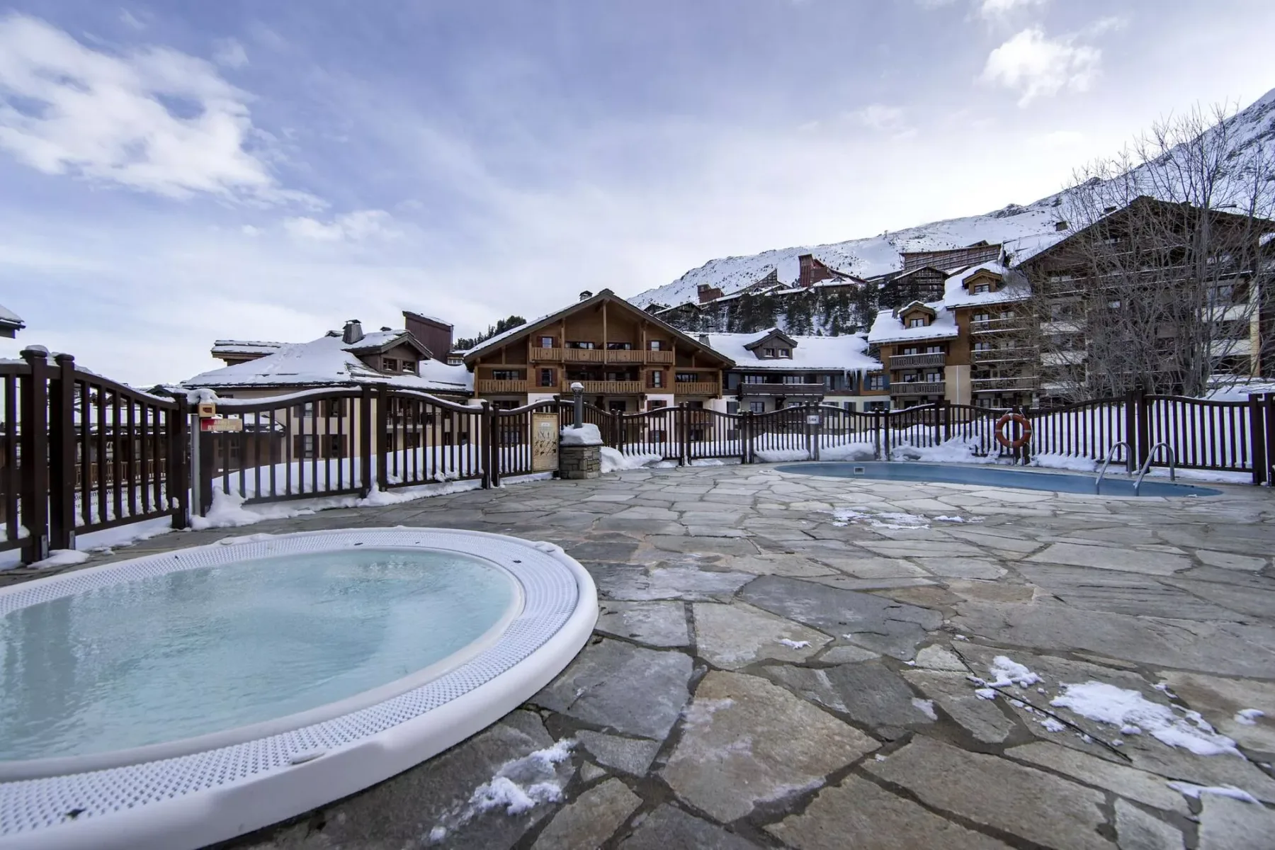 Una terraza exterior de piedra da a una pequeña piscina y edificios de montaña cubiertos de nieve.