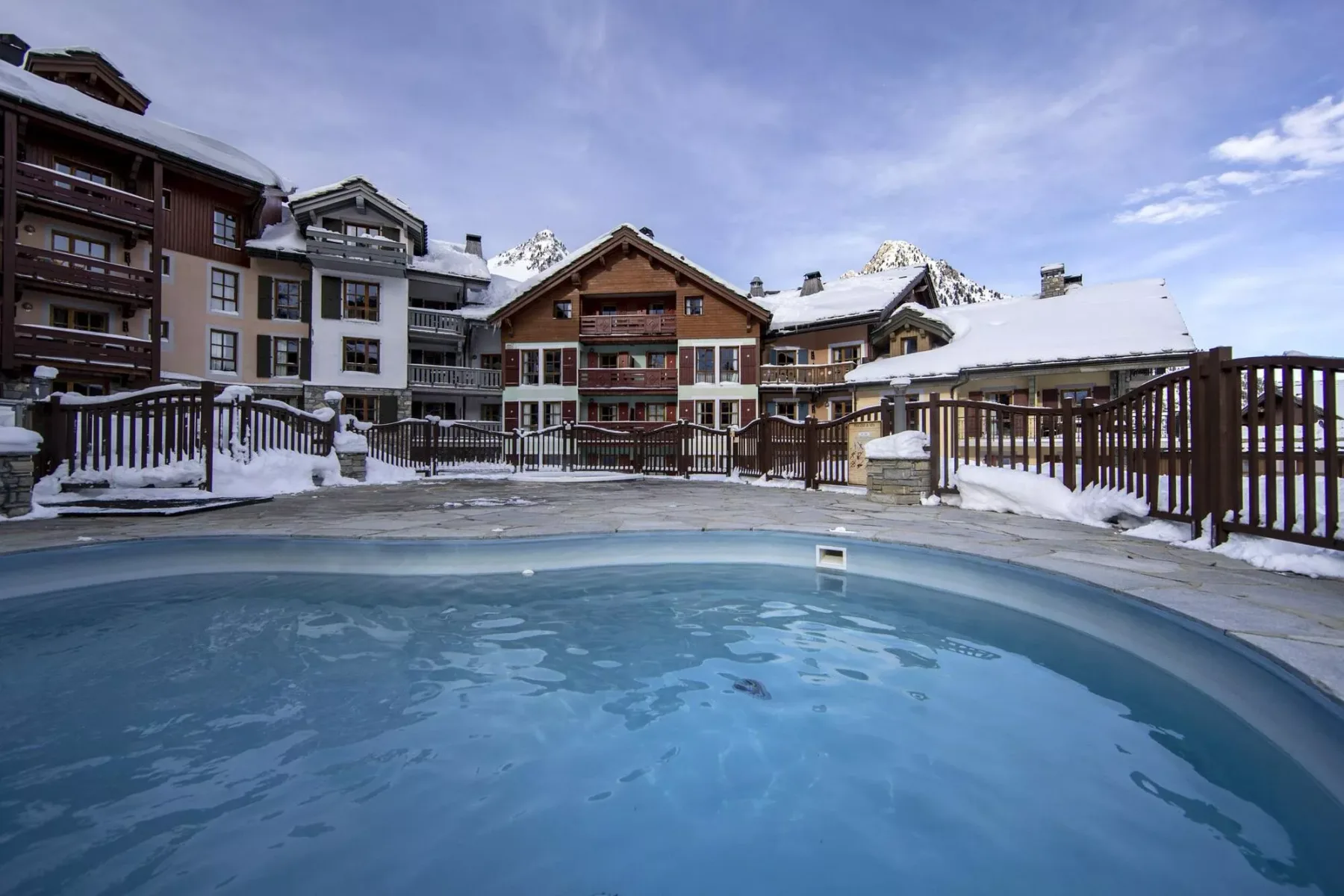 Una piscina al aire libre se encuentra frente a edificios de montaña cubiertos de nieve.