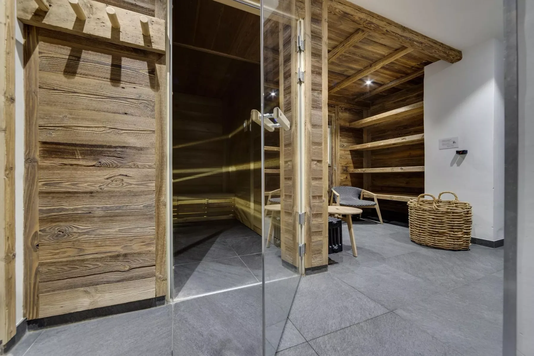 Una sauna revestida de madera da a una sala con suelo de baldosas, asientos y una cesta tejida.