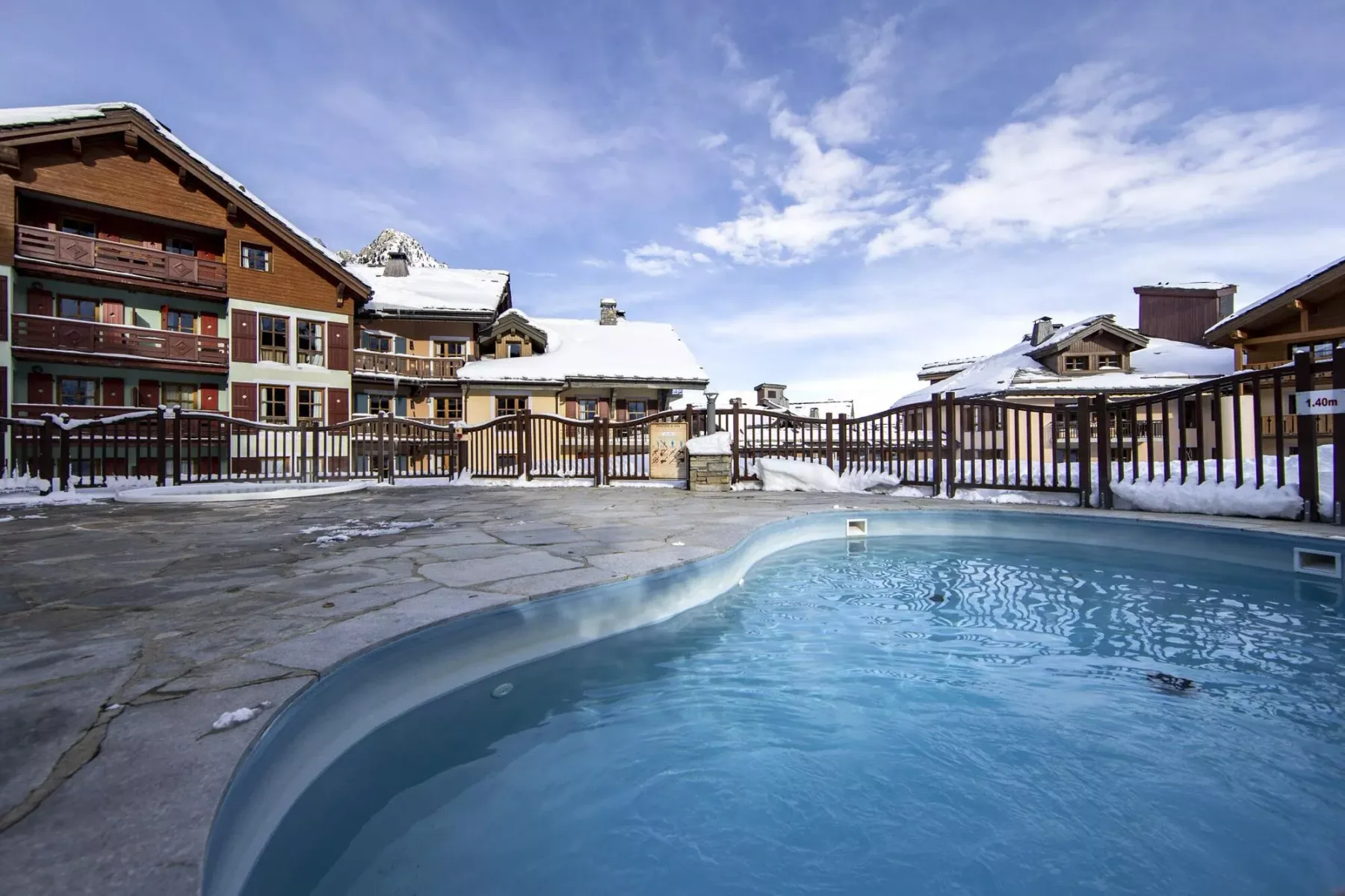 Una piscina al aire libre está junto a edificios cubiertos de nieve bajo un cielo parcialmente nublado.